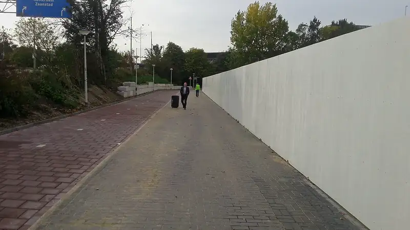 A wide city sidewalk and red brick bike path running beside a long white wall under a gray sky.
