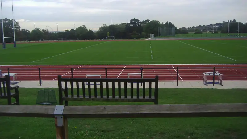 A red running track beside a green sports field, seen past a wooden bench and rail.