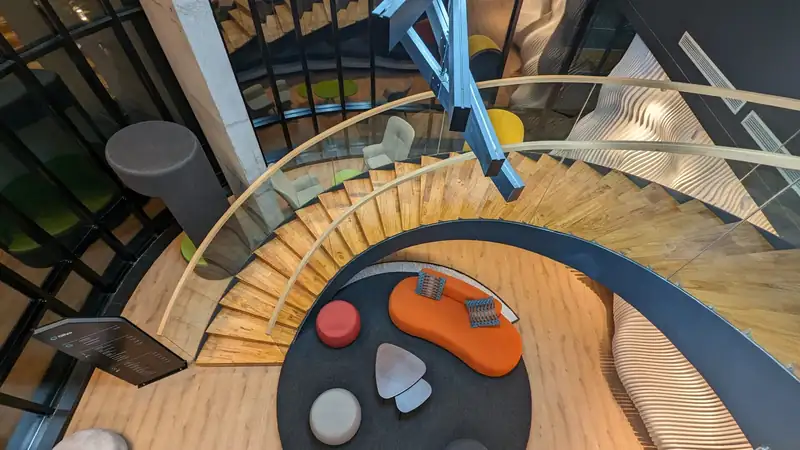 A curving wooden office staircase above a lounge with colorful seating, seen from above.