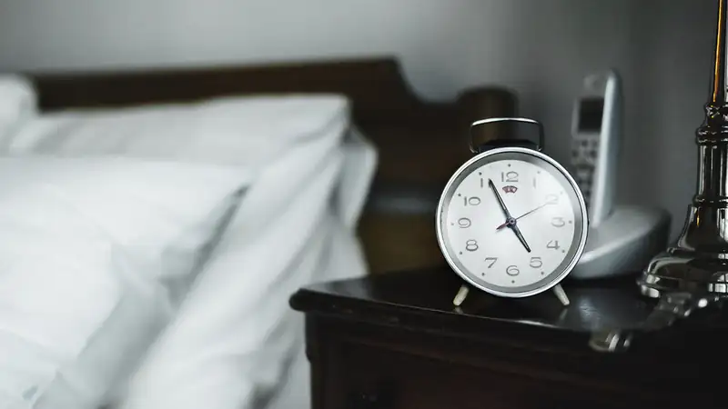 An analog alarm clock on a dark nightstand beside white pillows.