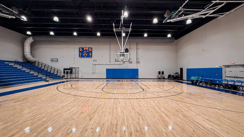 An empty indoor basketball court with a hardwood floor and blue bleachers under bright lights.