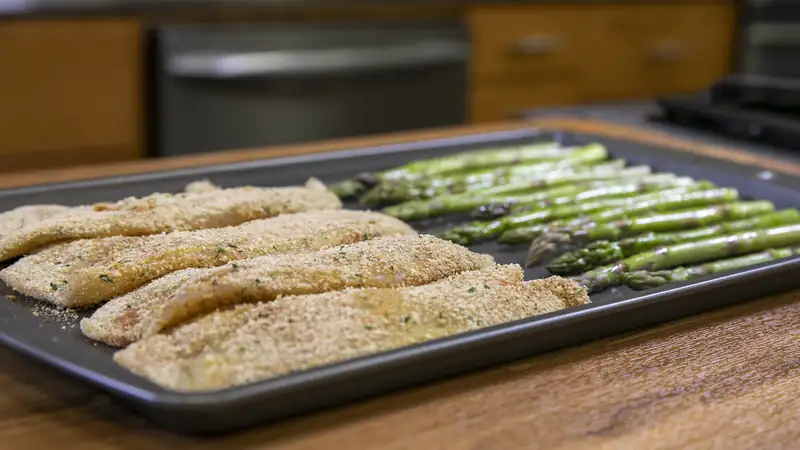 A sheet pan of breaded fish fillets and asparagus on a kitchen counter.